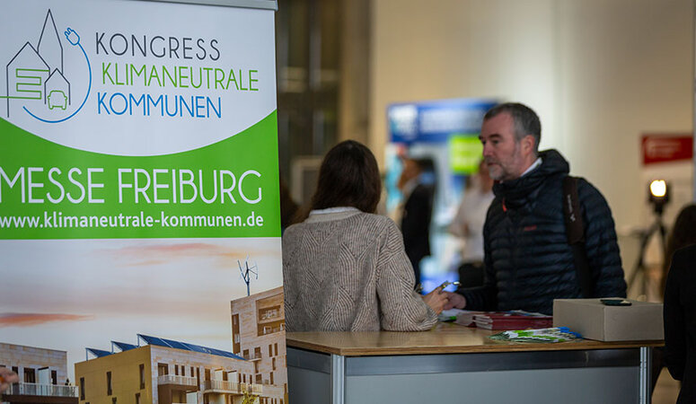 Im Vordergrund ein Aufsteller mit der Aufschrift "Kongress klimaneutrale Kommunen Messe Freiburg", dahinter 2 Personen im Gespräch