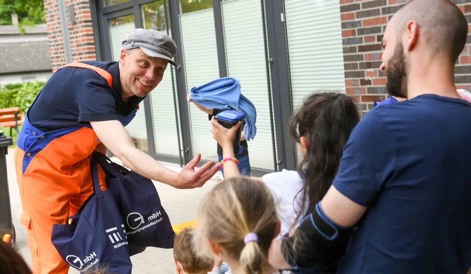 Ein Mann mit orangefarbener Latzhose – Herr Stinknich – im Gespräch mit Kindern (die nur von hinten zu sehen sind)
