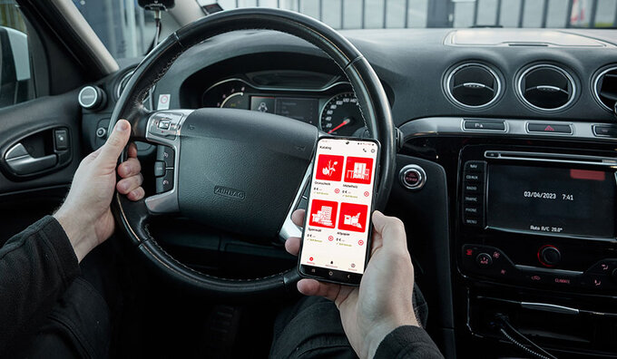 Blick von der Rückbank eines PKWs Richtung Fahrer; von diesem sieht man nur die Hände; in der rechten Hand ein Handy mit der MAEX-App auf dem Display